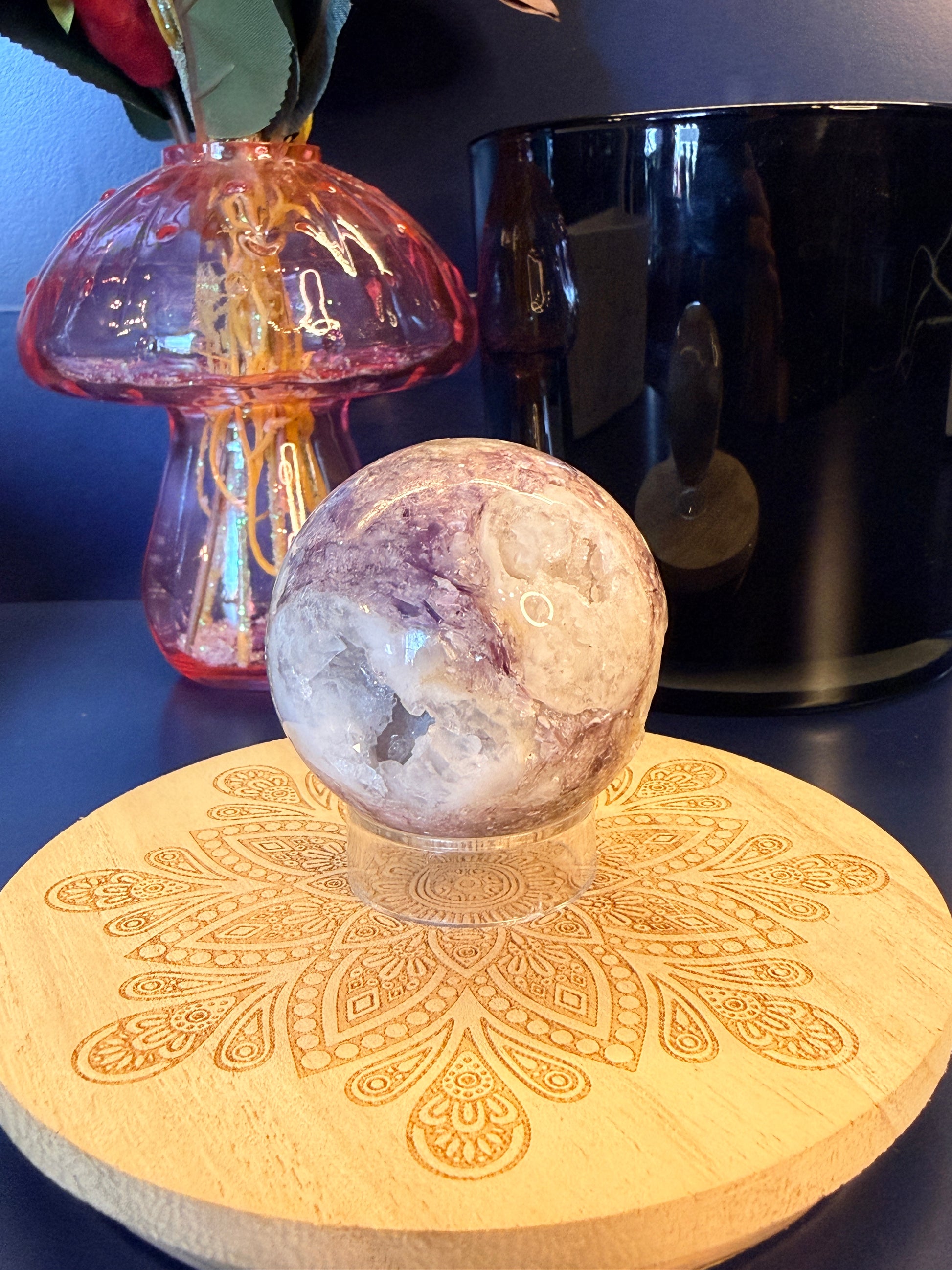 A white and purple crystal sphere sitting on a decorative wooden coaster in front of a glass mushroom and black jar