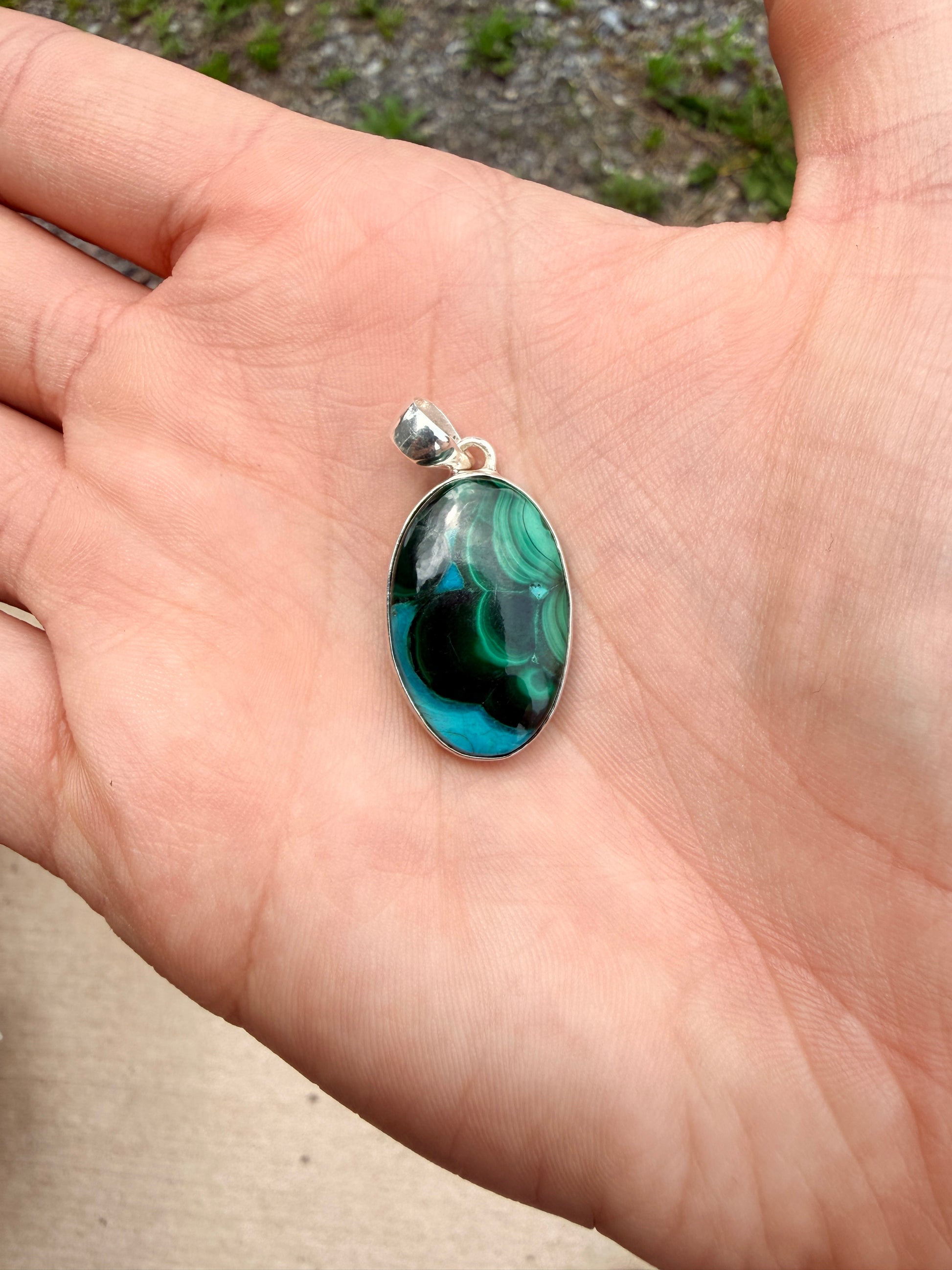 Hand holding a green and blue malachite and chrysocola sterling silver pendant in front of a blurred nature background
