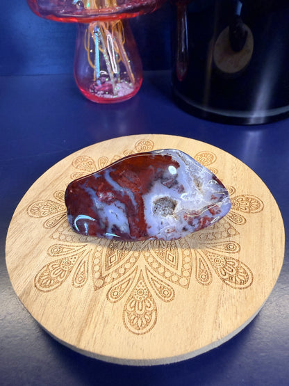Red and light blue mexican agate freeform on a decorative wooden coaster in front of a mushroom and black jar