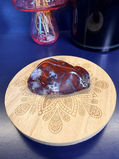 Red and light blue mexican agate freeform on a decorative wooden coaster in front of a mushroom and black jar