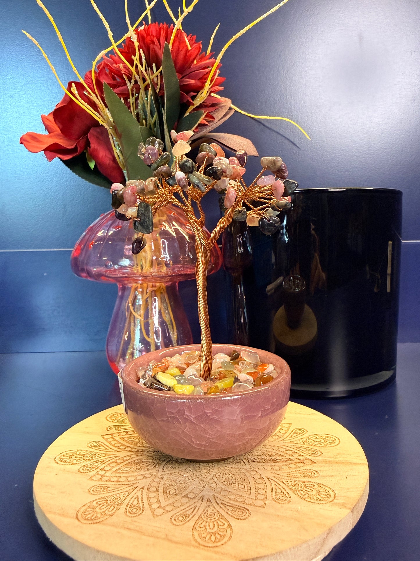 A multi-colored crystal chip tree made of copper wire in a pink bowl on a wooden coaster in front of a pink mushroom and black jar