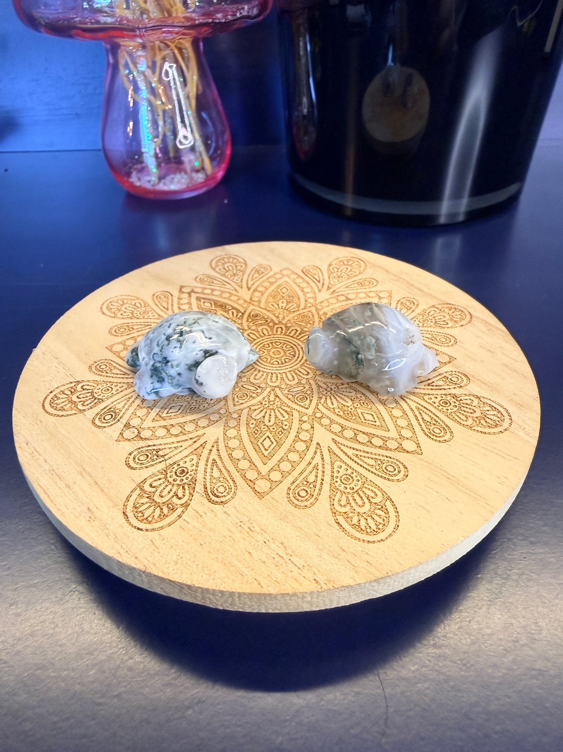 Two green and white crystal turtles on a decorative wooden coaster in front of a vase and black jar