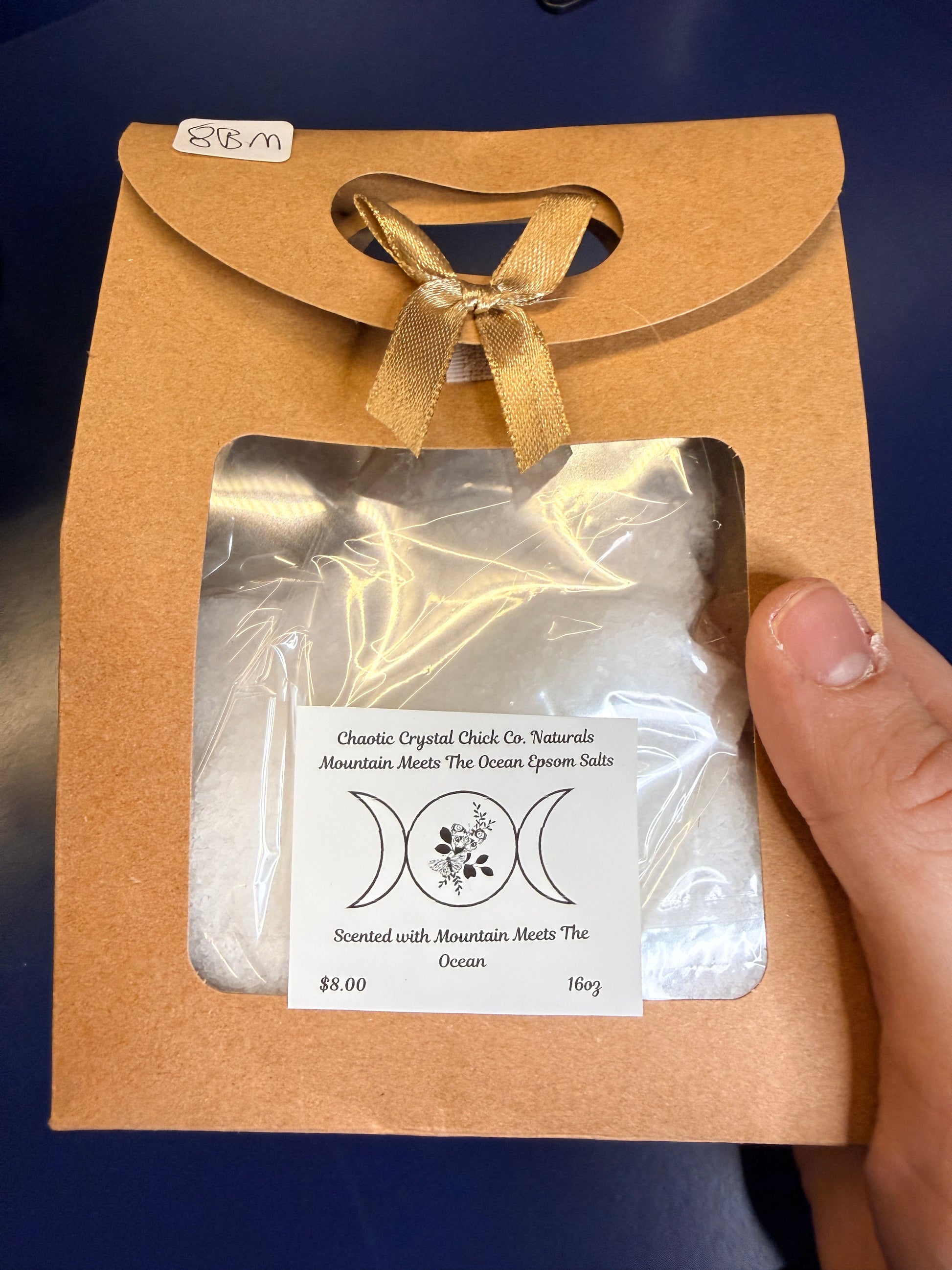 Hand holding white epsom salt in a brown paper container with a bow, on a wooden coaster