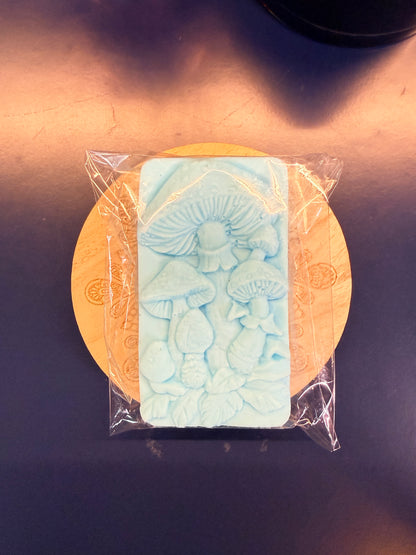 Blue soap bar with decorative design on a wooden coaster, wrapped in clear plastic on a dark surface.