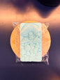 Blue soap bar with decorative design on a wooden coaster, wrapped in clear plastic on a dark surface.