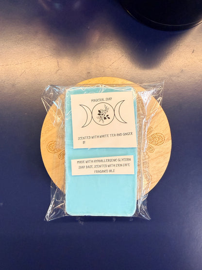 Blue soap bar with two labels on a wooden coaster, wrapped in clear plastic on a dark surface.