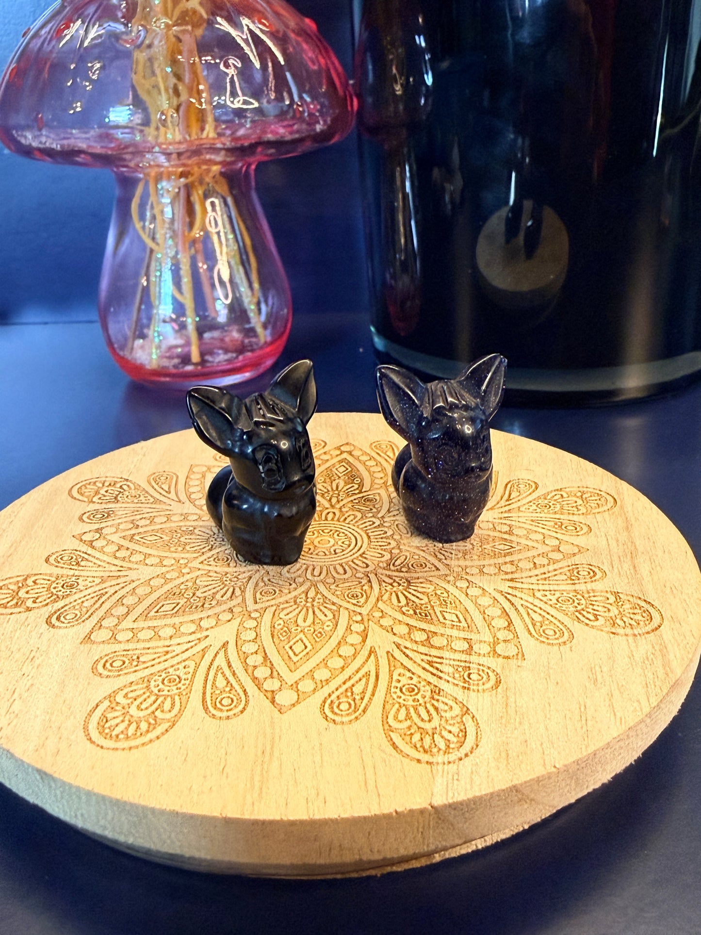 Two eevee crystal shapes, obsidian and blue goldstone,  sitting on a coaster in front of a glass mushroom and black jar