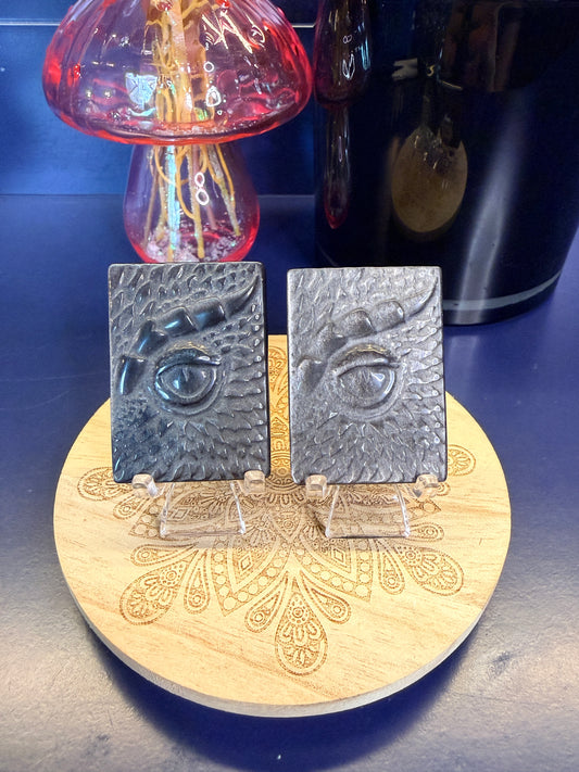 Two black and silver obsidian dragon eye slabs on a decorative wooden coaster in front of a pink mushroom and black jar