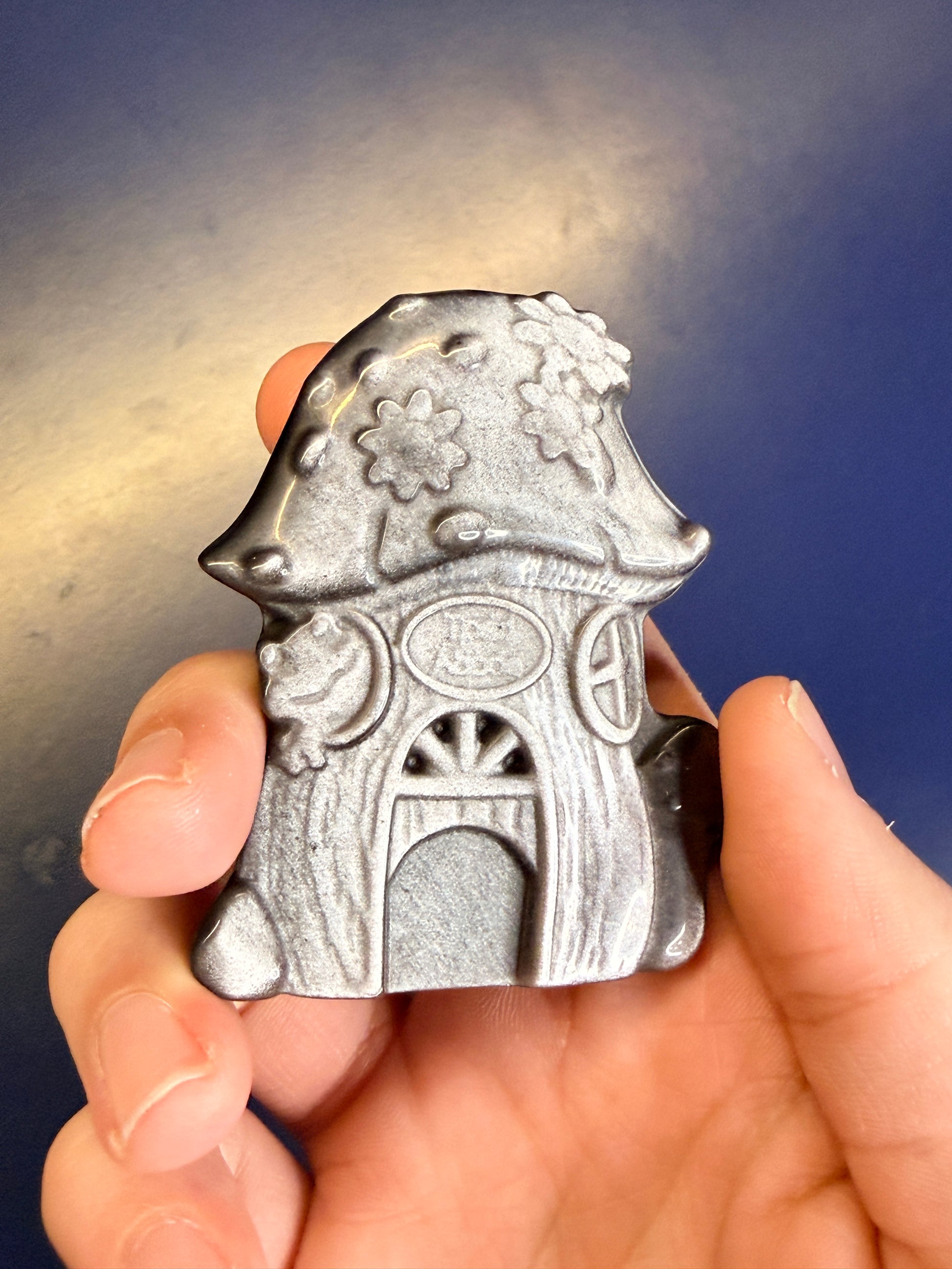 Hand holding a silver and black obsidian mushroom house on a blue background