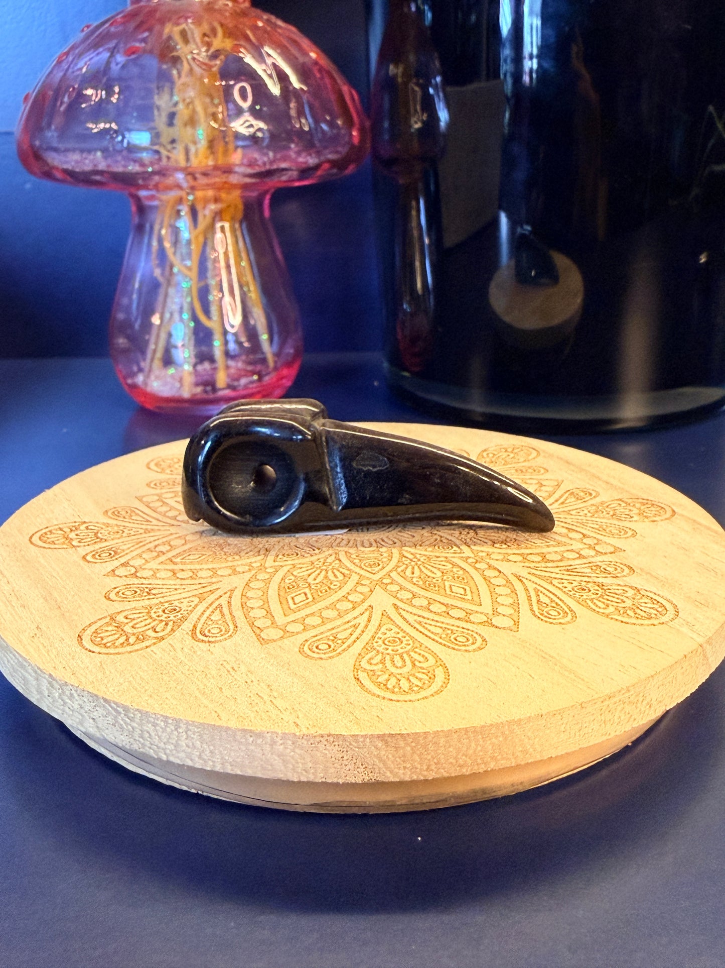 A black obsidian raven skull crystal sitting on a decorative wooden coaster in front of a glass mushroom and black jar