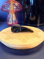 A black obsidian raven skull crystal sitting on a decorative wooden coaster in front of a glass mushroom and black jar