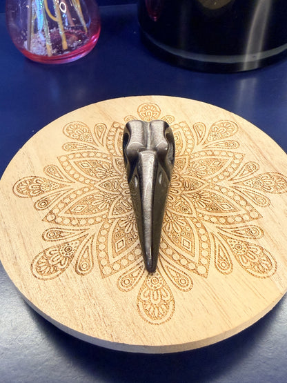 A black obsidian raven skull crystal sitting on a decorative wooden coaster in front of a glass mushroom and black jar
