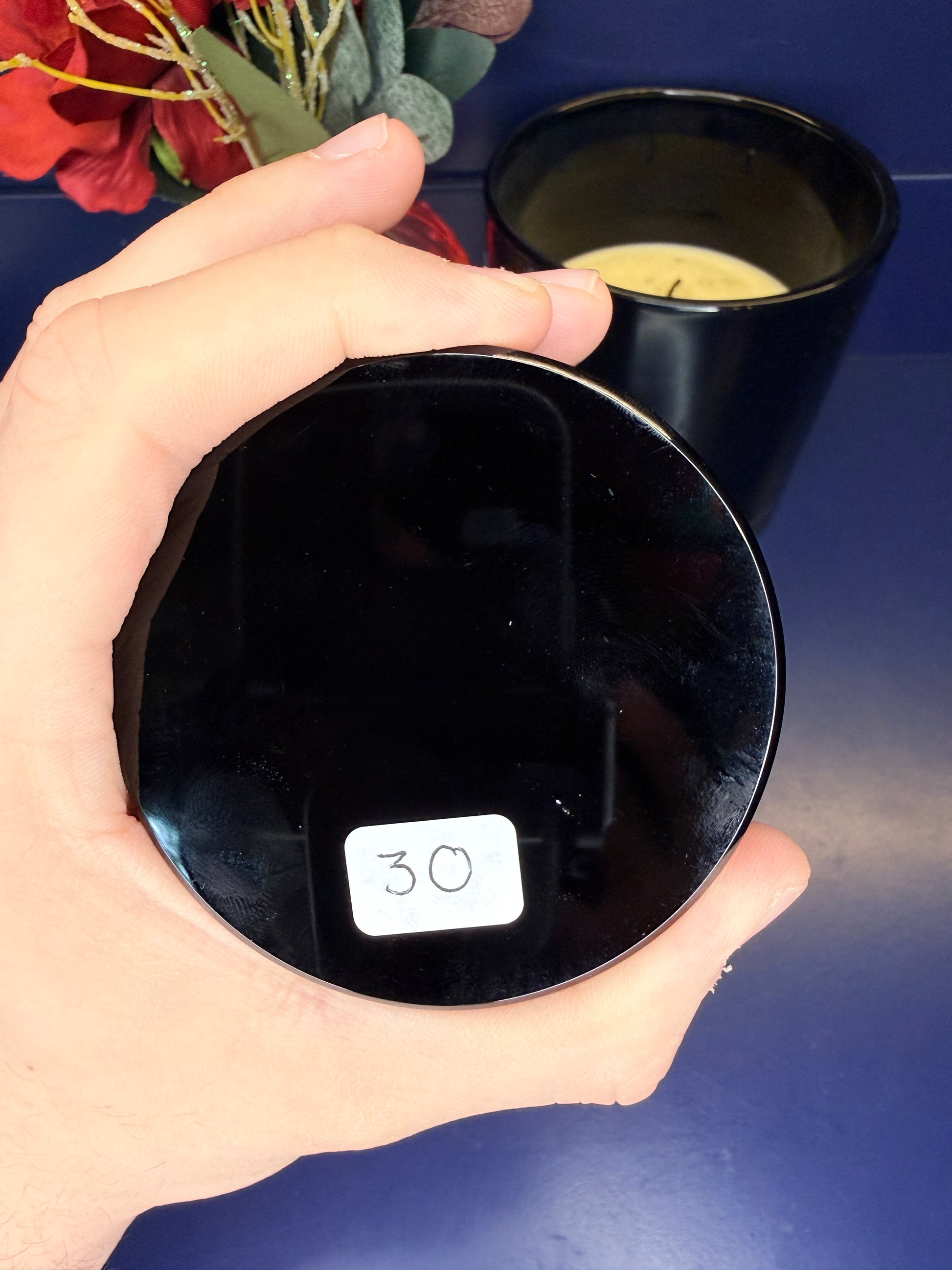 Hand holding a round black obsidian scrying mirror in front of a red flower and black candle jar