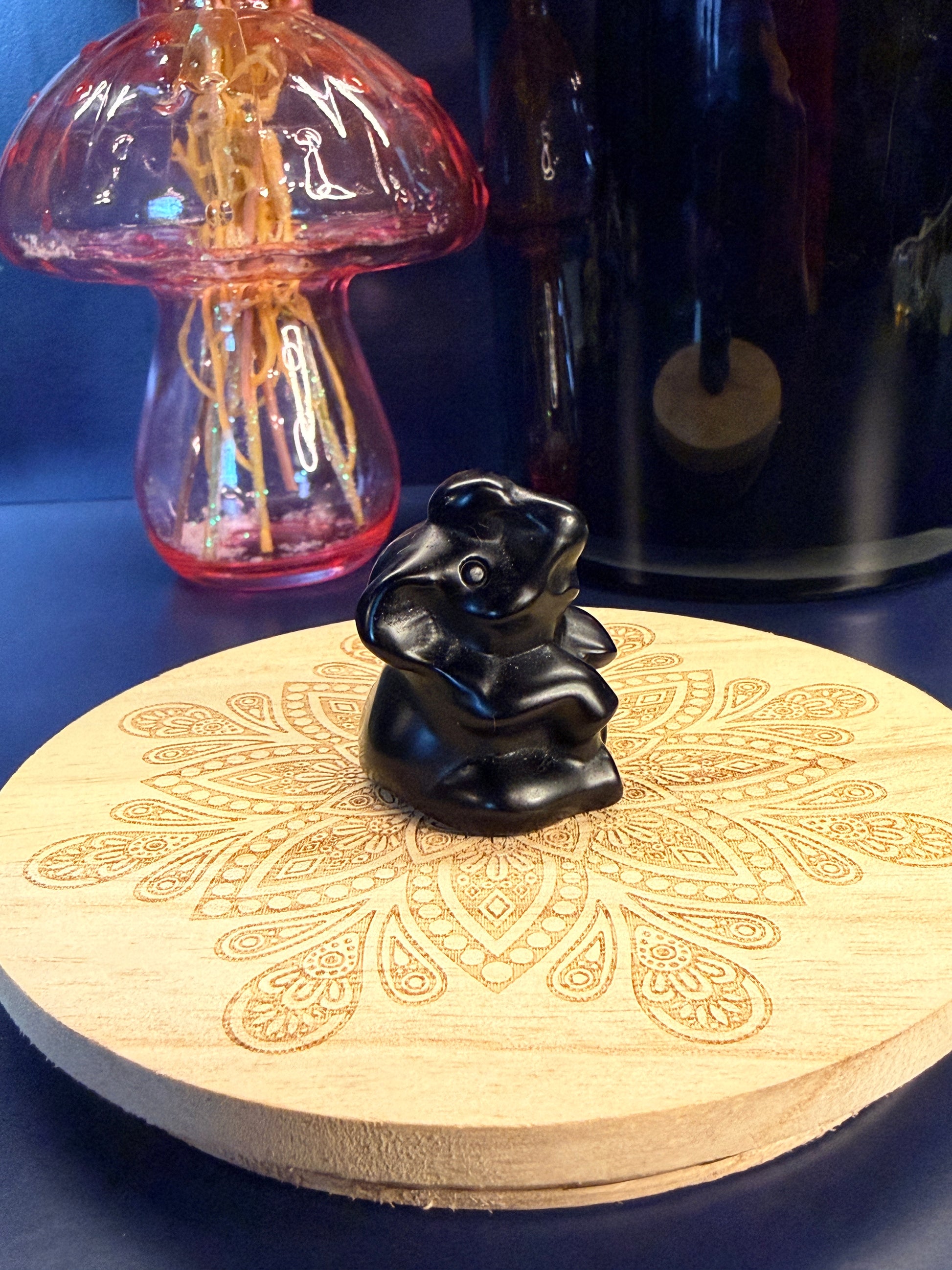 An obsidian sitting elephant crystal sitting on a decorative wooden coaster in front of a glass mushroom and black jar
