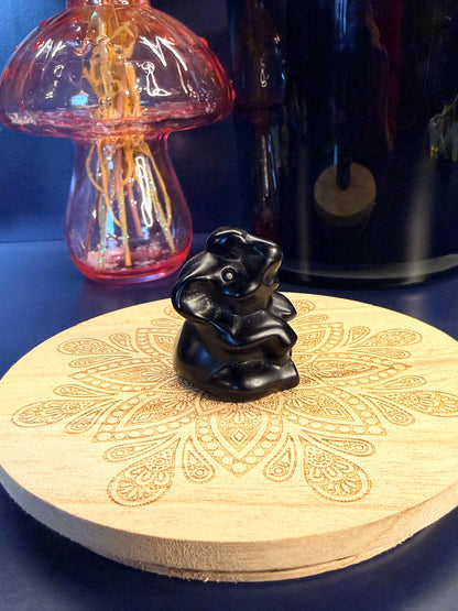 An obsidian sitting elephant crystal sitting on a decorative wooden coaster in front of a glass mushroom and black jar