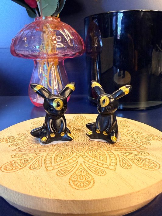 Two black and gold crystal umbreons sitting on a decorative wooden coaster in front of a glass mushroom and black jar
