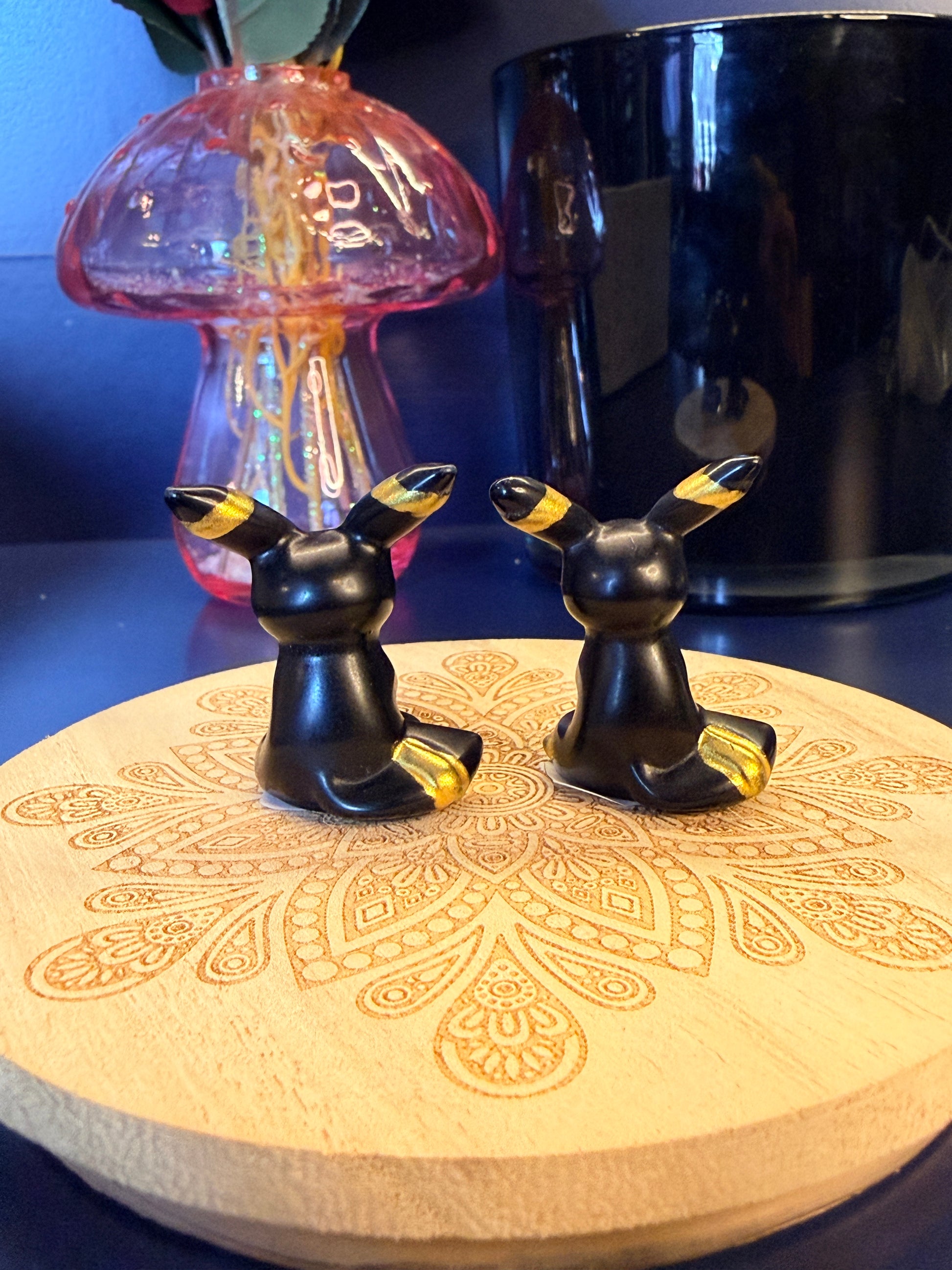 Two black and gold crystal umbreons sitting on a decorative wooden coaster in front of a glass mushroom and black jar