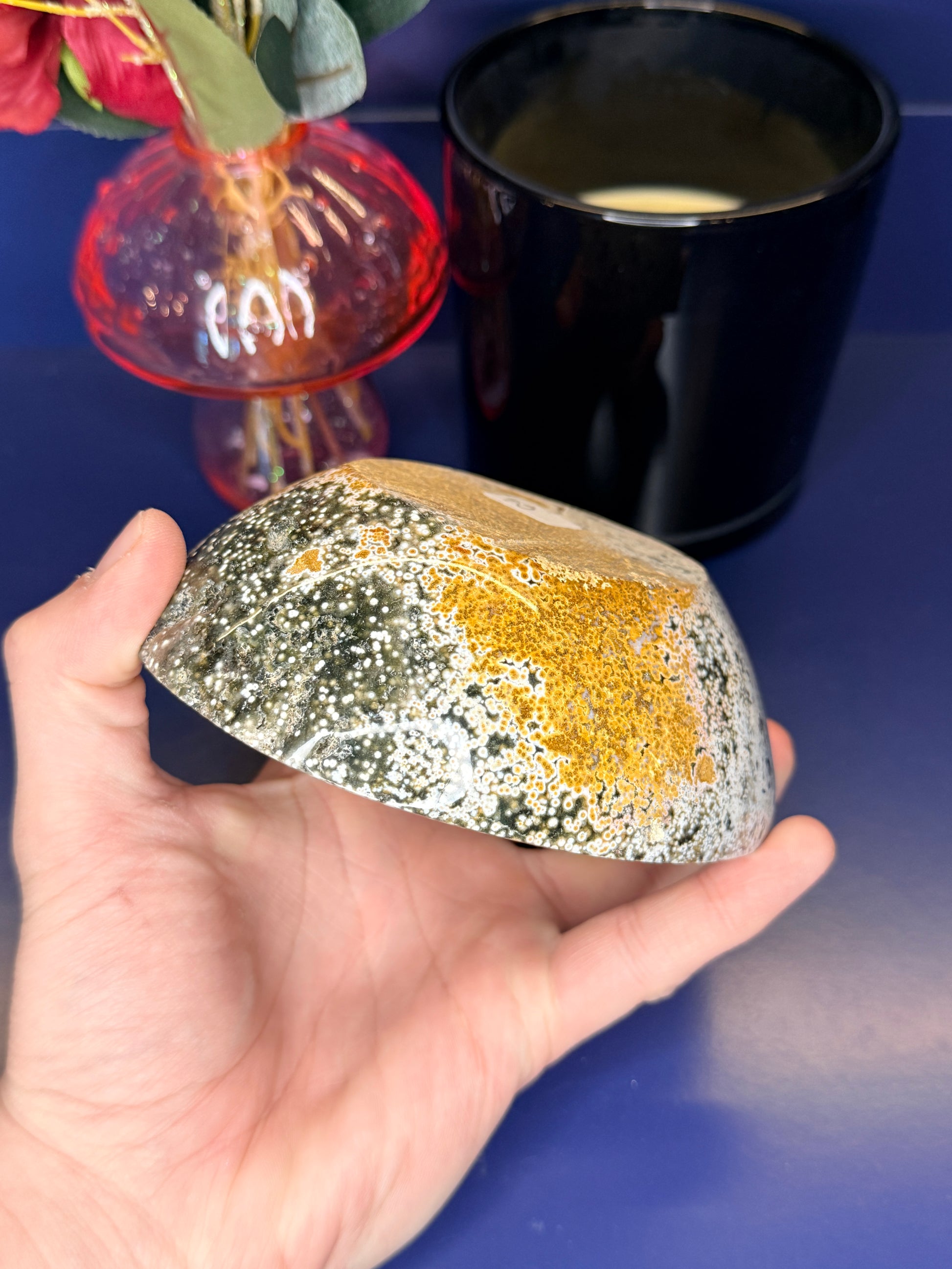 Hand holding white, green, and yellow ocean jasper bowl in front of a pink mushroom vase and black jar on a blue background