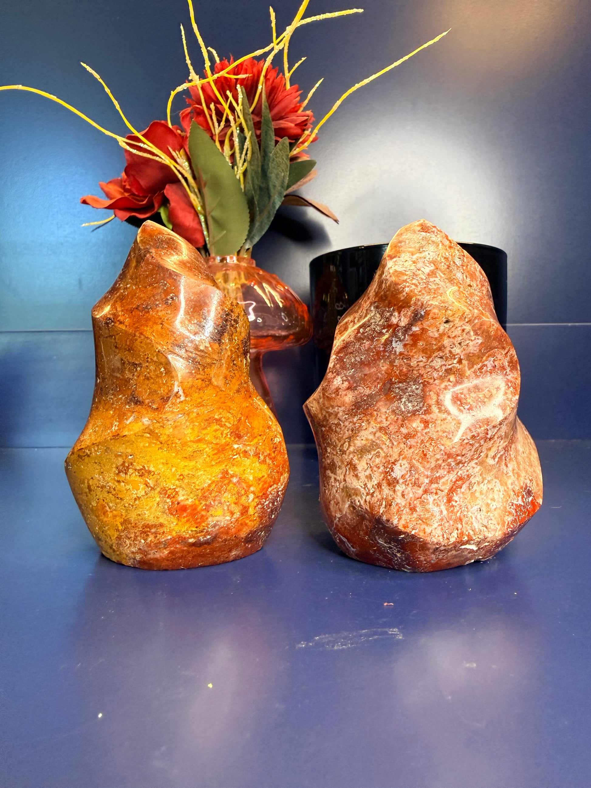 Two orange and red ocean jasper flames in front of a red flower and black jar on a blue background