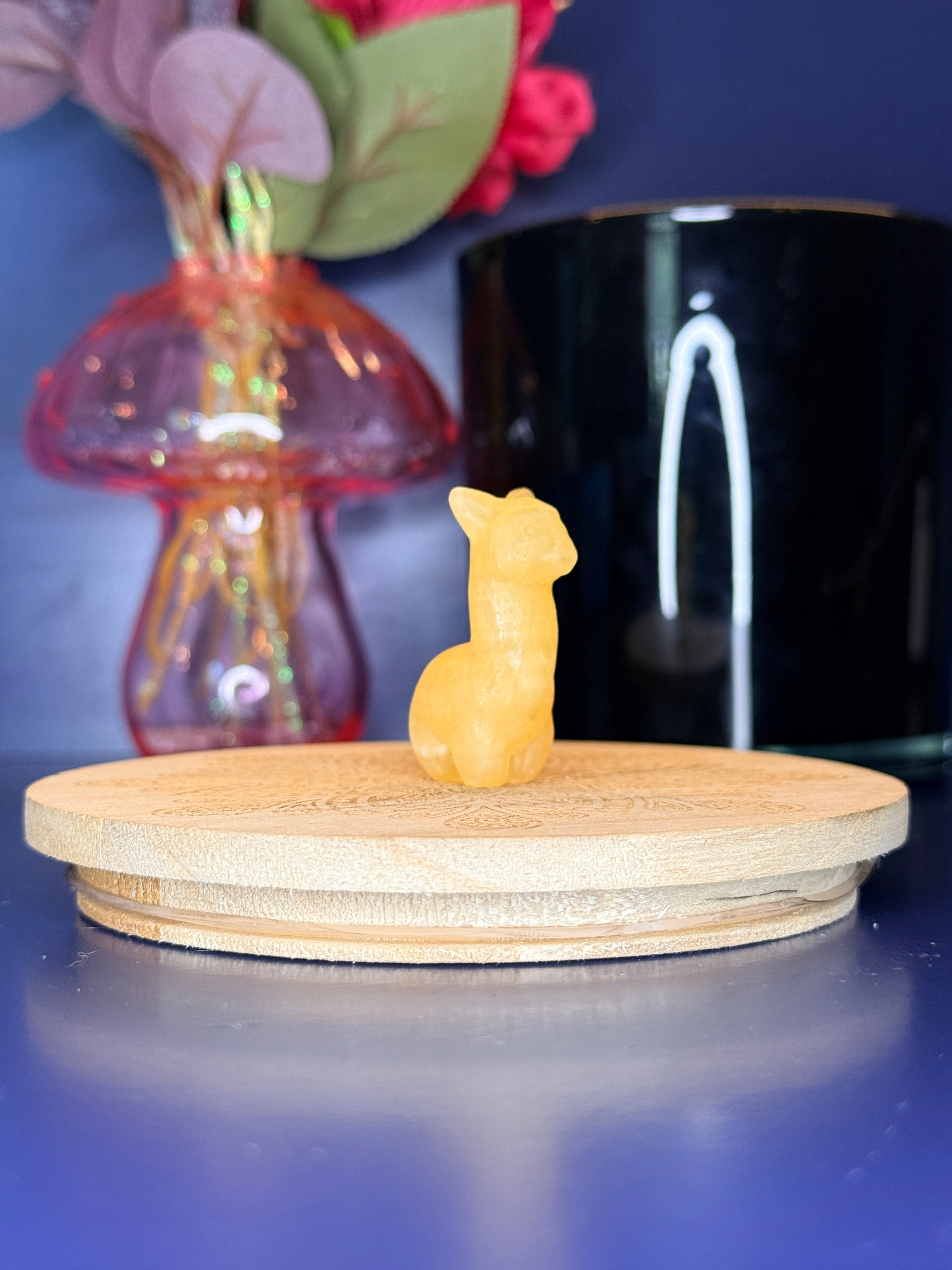 Orange calcite alpaca carving in front of a pink vase and black jar on a wooden coaster against a blue background
