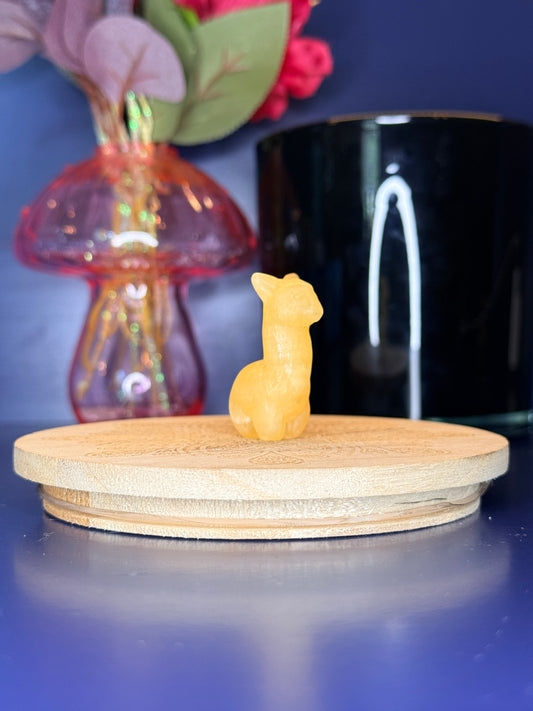 Orange calcite alpaca carving in front of a pink vase and black jar on a wooden coaster against a blue background