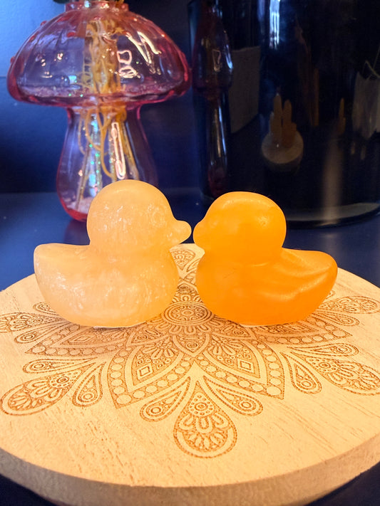 Two orange crystal ducks sitting on a decorative wooden coaster in front of a glass mushroom and black jar