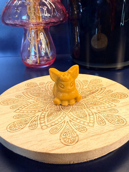 An orange calcite flareon crystal sitting on a decorative wooden coaster in front of a glass mushroom and black jar