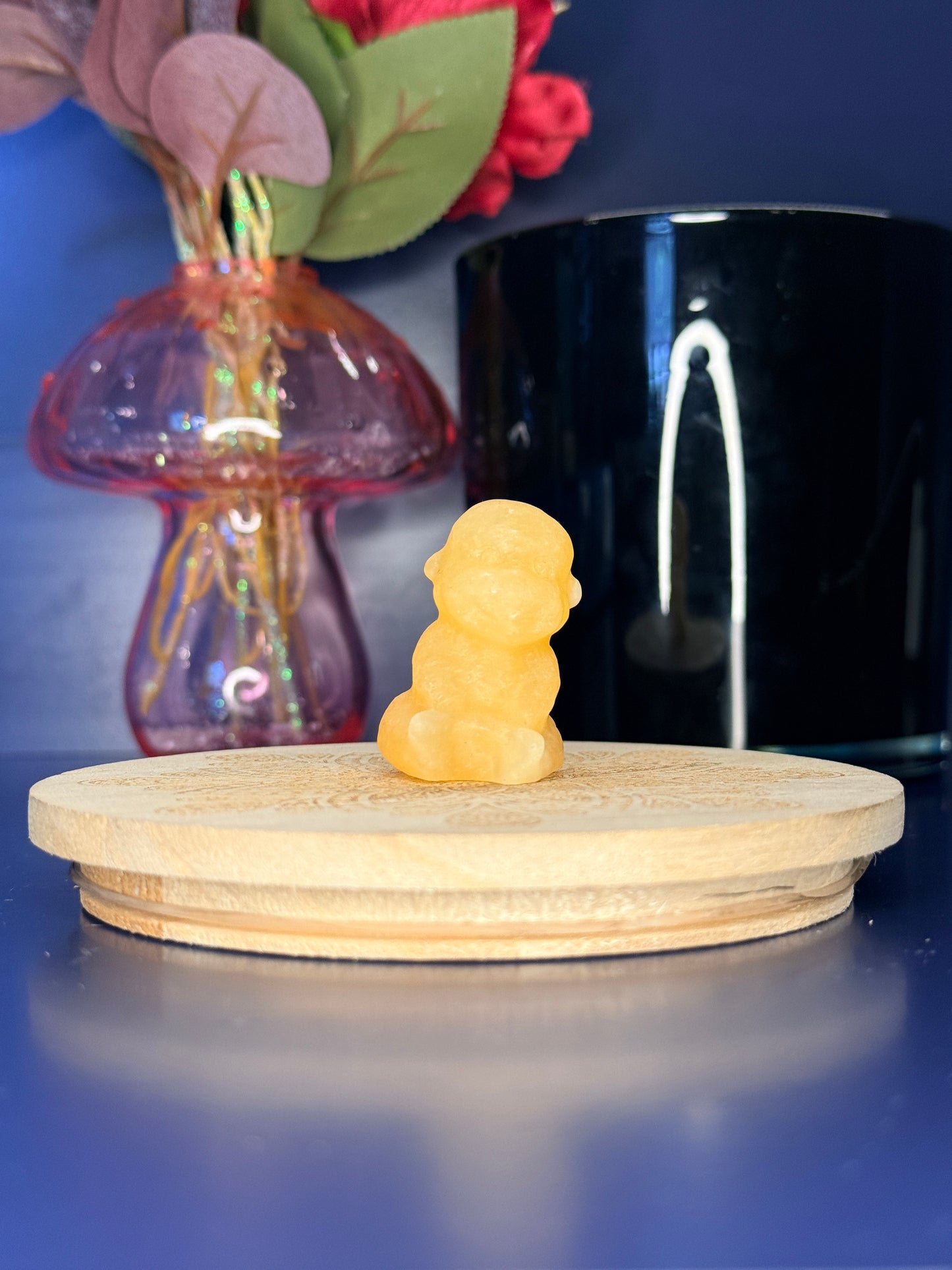Orange calcite monkey carving in front of a pink vase and black jar on a wooden coaster against a blue background