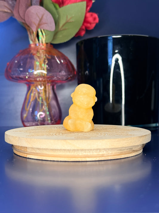 Orange calcite monkey carving in front of a pink vase and black jar on a wooden coaster against a blue background
