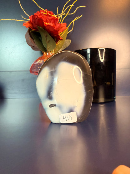 A white and grey orca agate freeform in front of a flower and black jar
