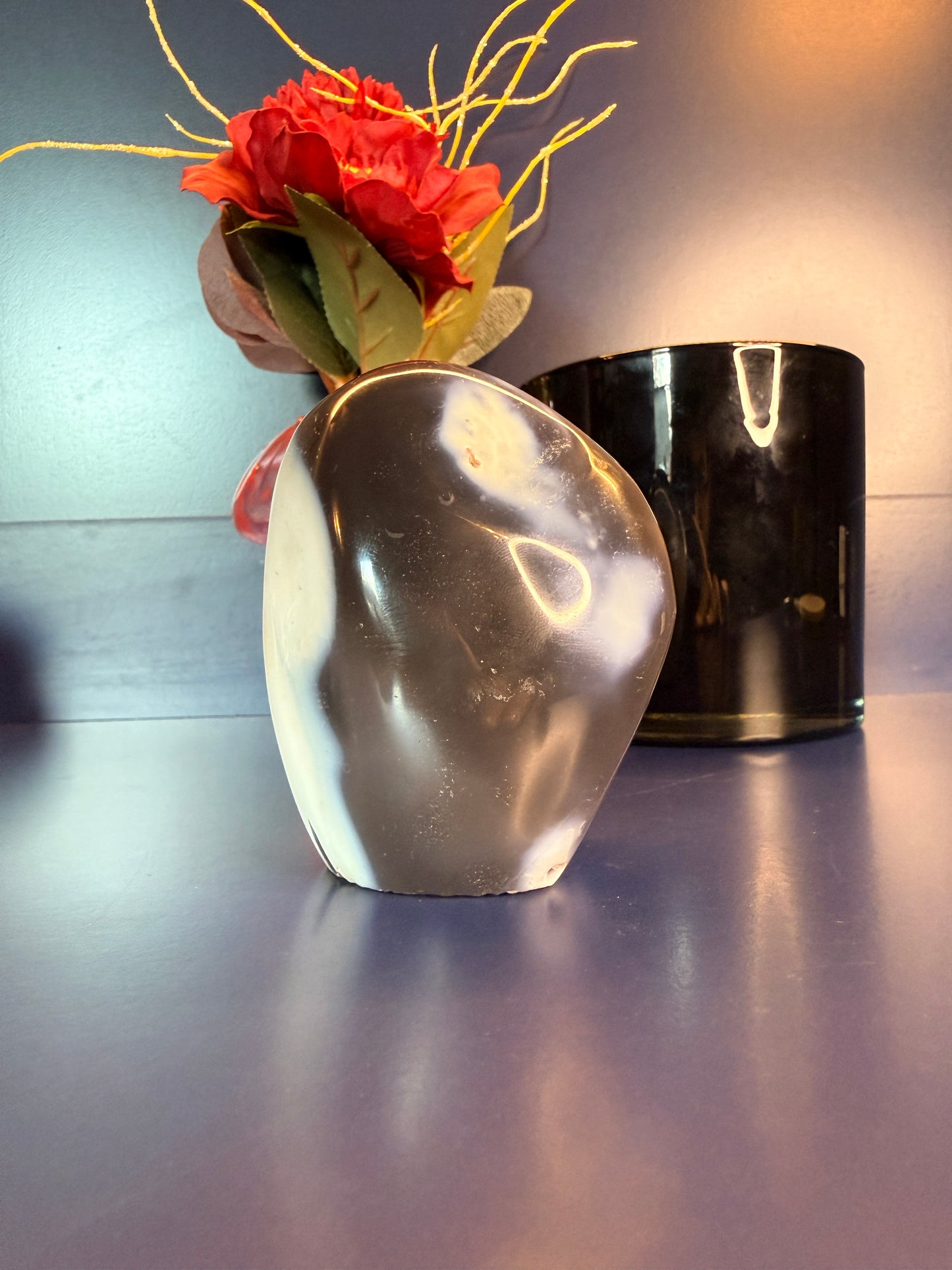 A white and grey orca agate freeform in front of a flower and black jar