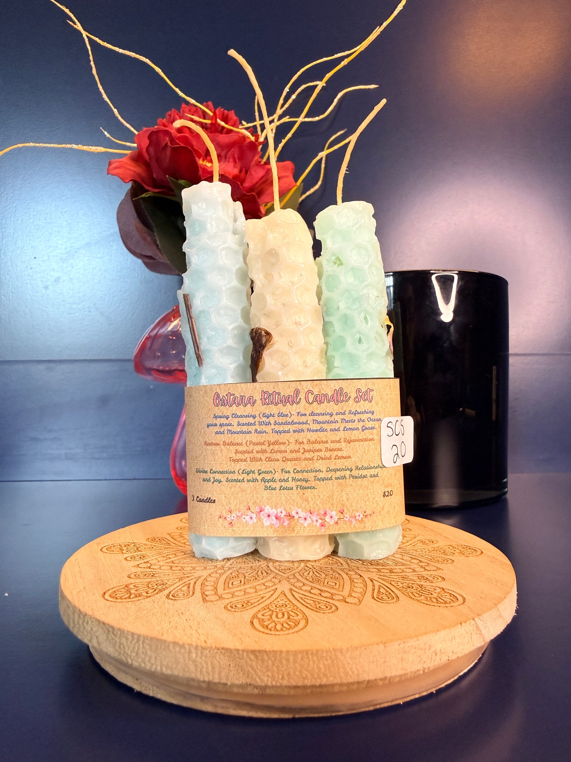 Three rolled candles with a label on a decorative wooden coaster in front of a red flower and black jar