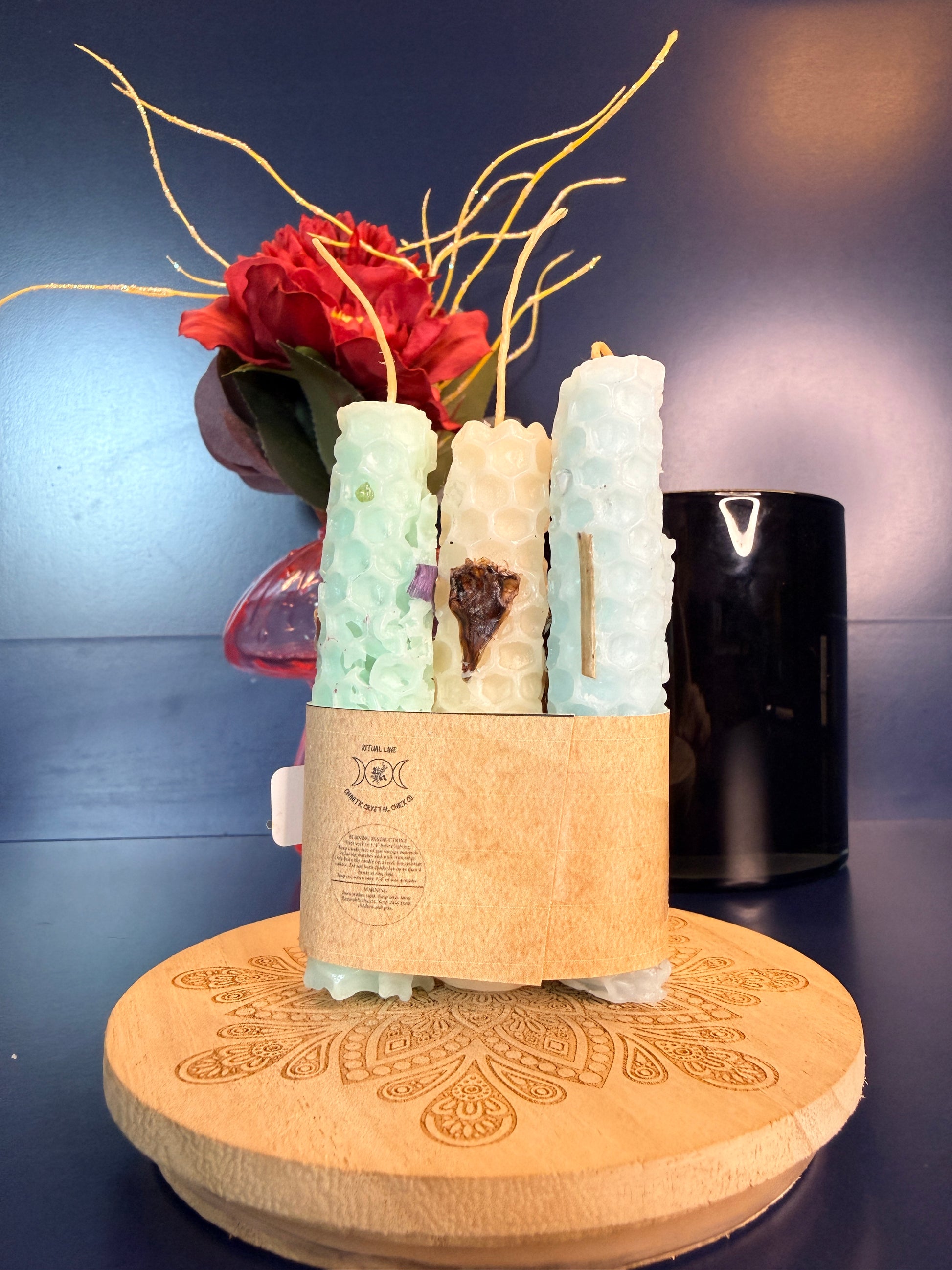Three rolled candles with a label on a decorative wooden coaster in front of a red flower and black jar