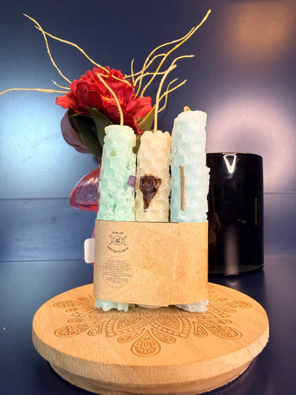 Three rolled candles with a label on a decorative wooden coaster in front of a red flower and black jar