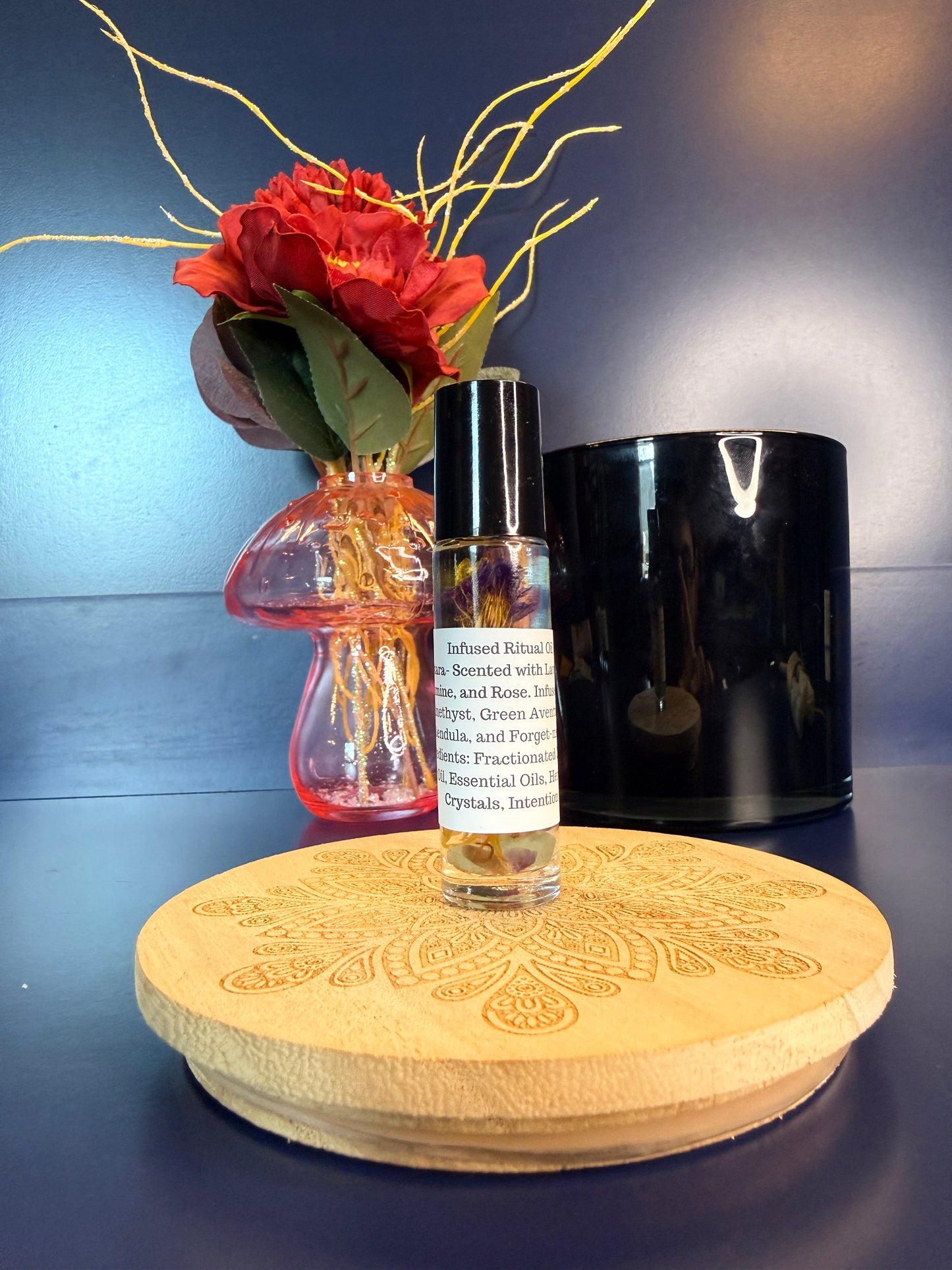Body Oil Vial sitting on a decorative wooden coaster in front of a red flower and black jar