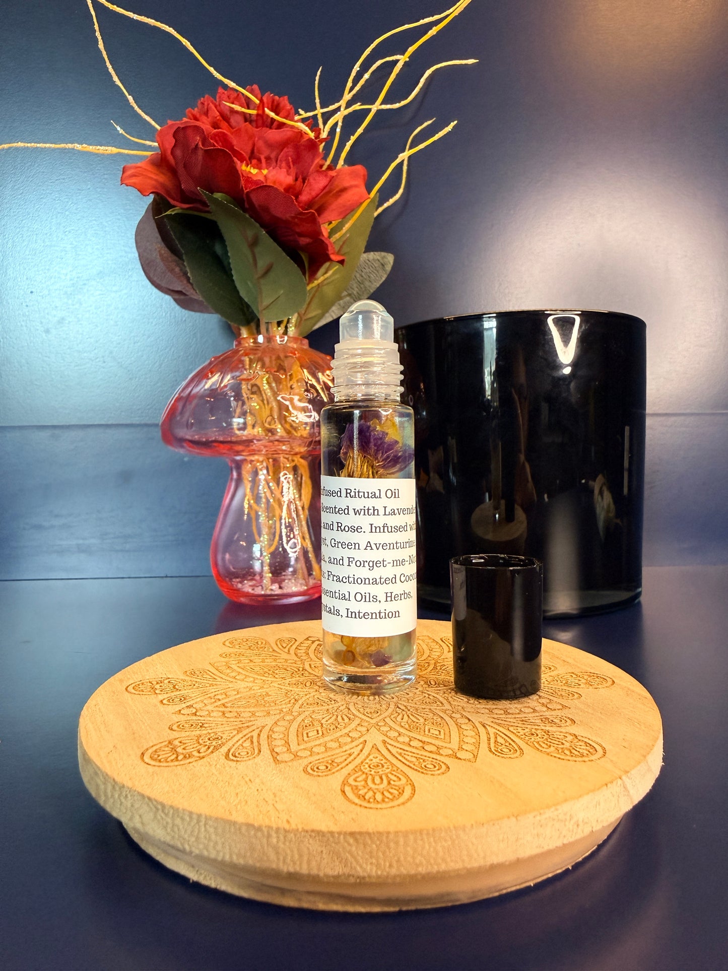 Body Oil Vial sitting on a decorative wooden coaster in front of a red flower and black jar