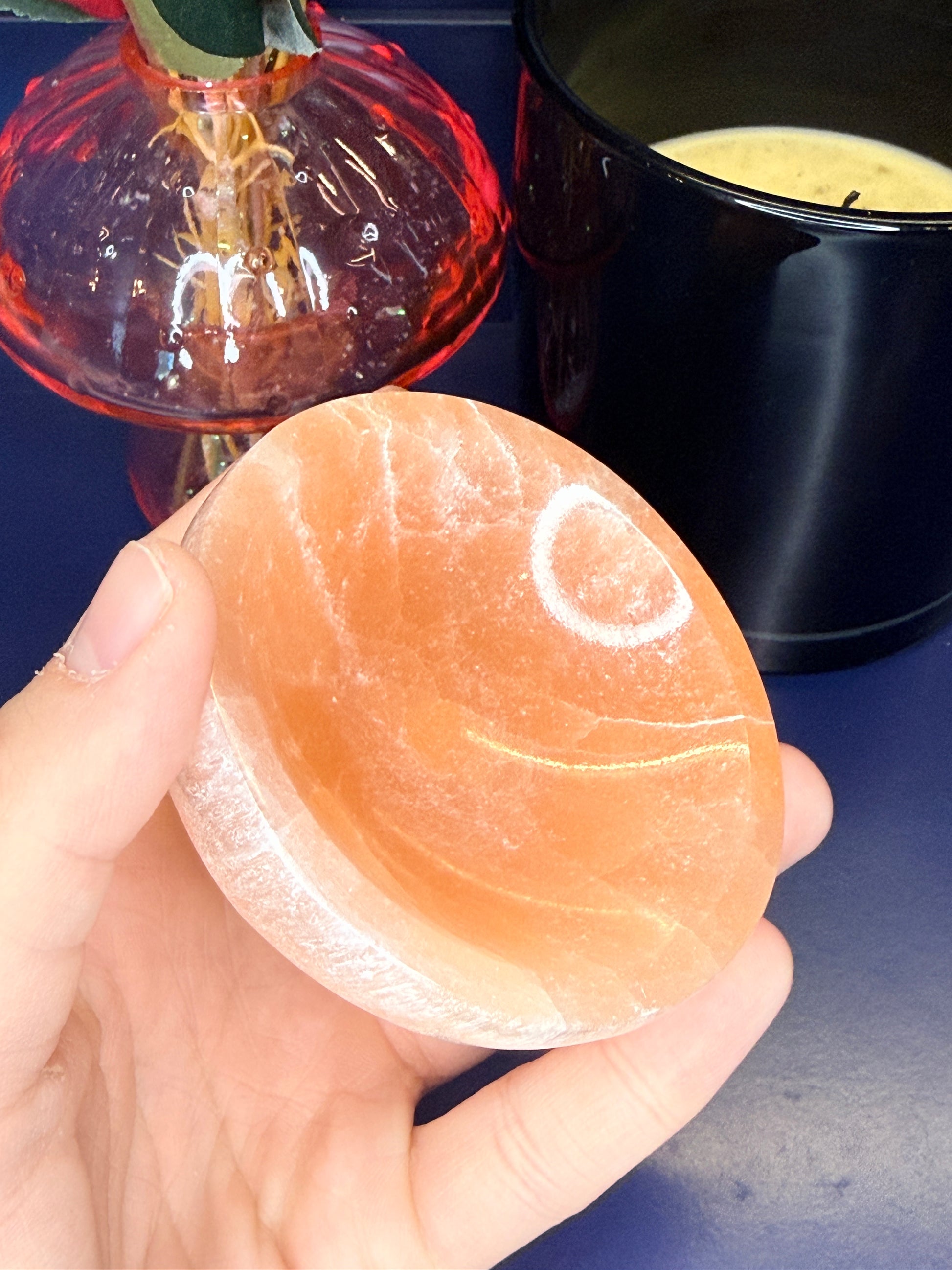 Hand holding a round peach selenite bowl in front of a pink mushroom vase and black candle jar on a blue background
