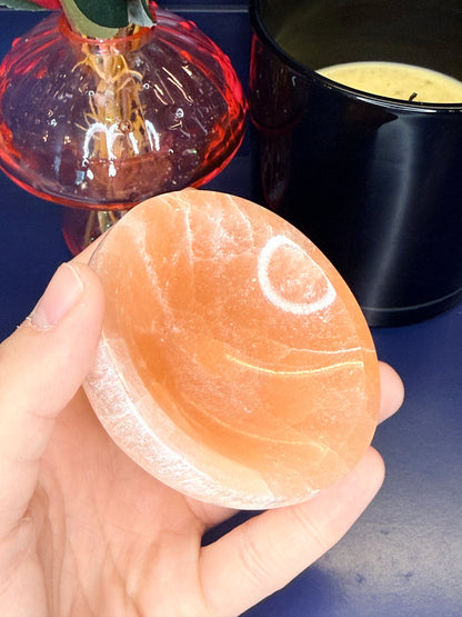 Hand holding a round peach selenite bowl in front of a pink mushroom vase and black candle jar on a blue background