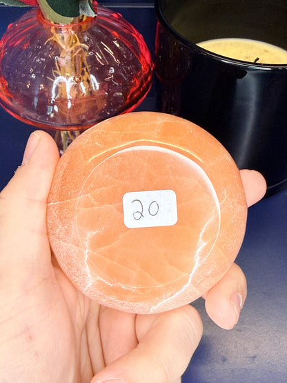 Hand holding a round peach selenite bowl with a label in front of a pink mushroom vase and black candle jar on a blue background