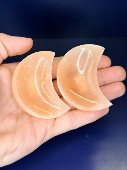 Hand holding two peach selenite moon bowls on a blue background