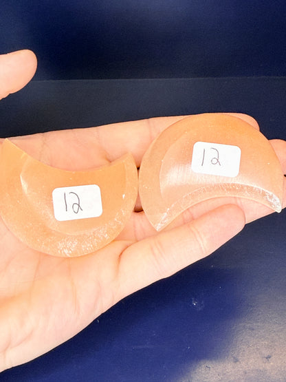 Hand holding two peach selenite moon bowls with labels on a blue background