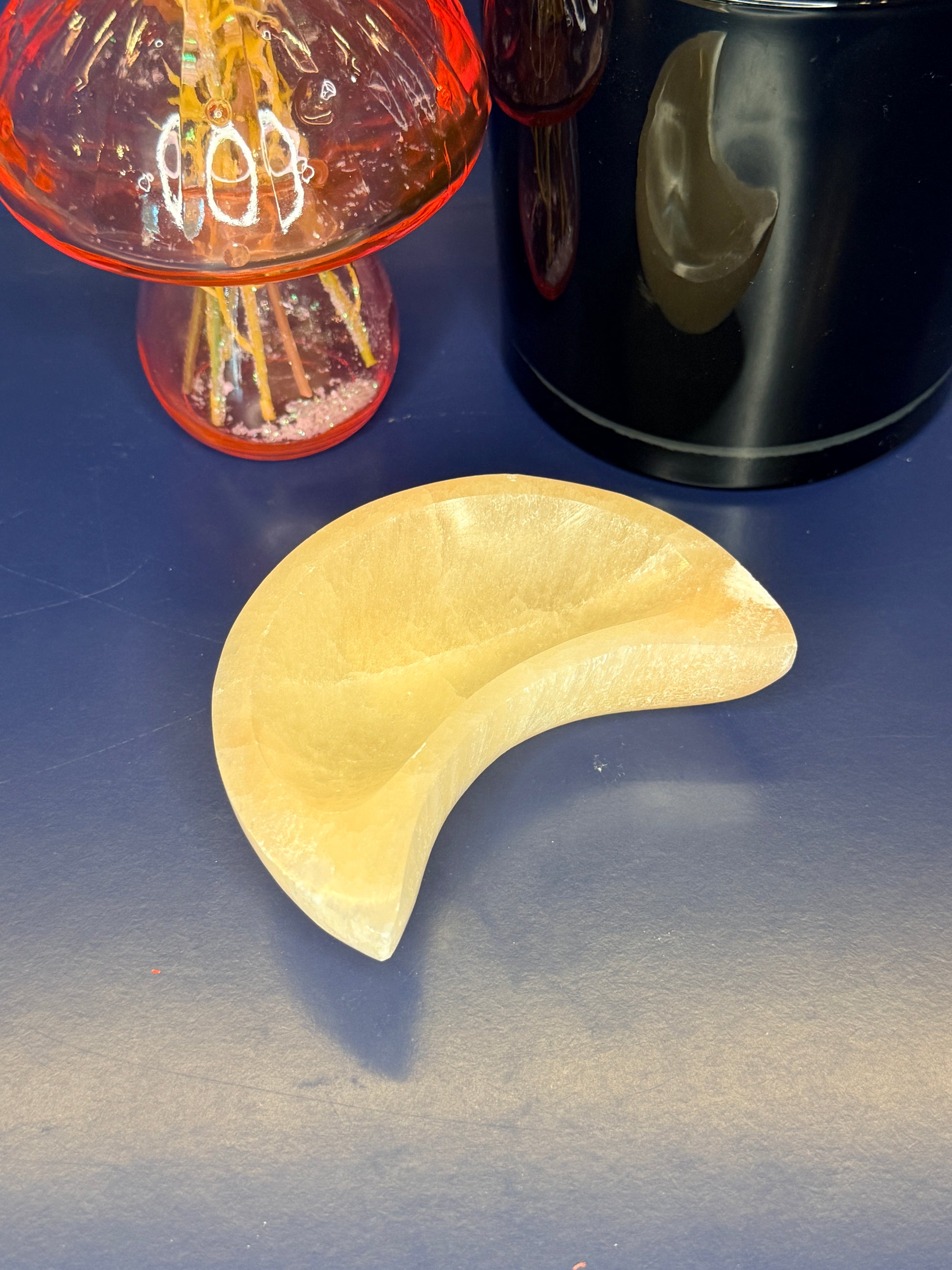 A peach selenite moon bowl sitting in front of a pink mushroom vase and black jar on a blue background