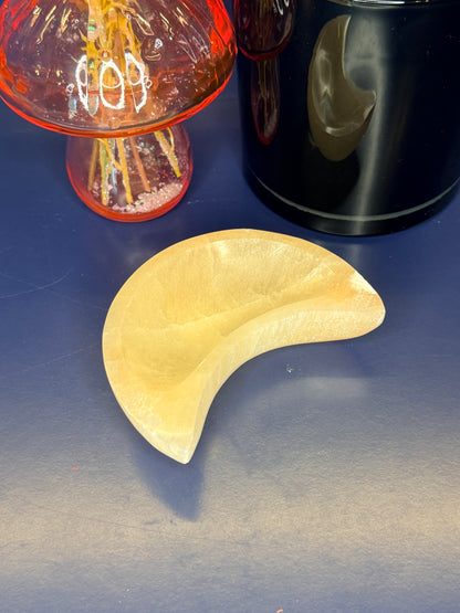 A peach selenite moon bowl sitting in front of a pink mushroom vase and black jar on a blue background