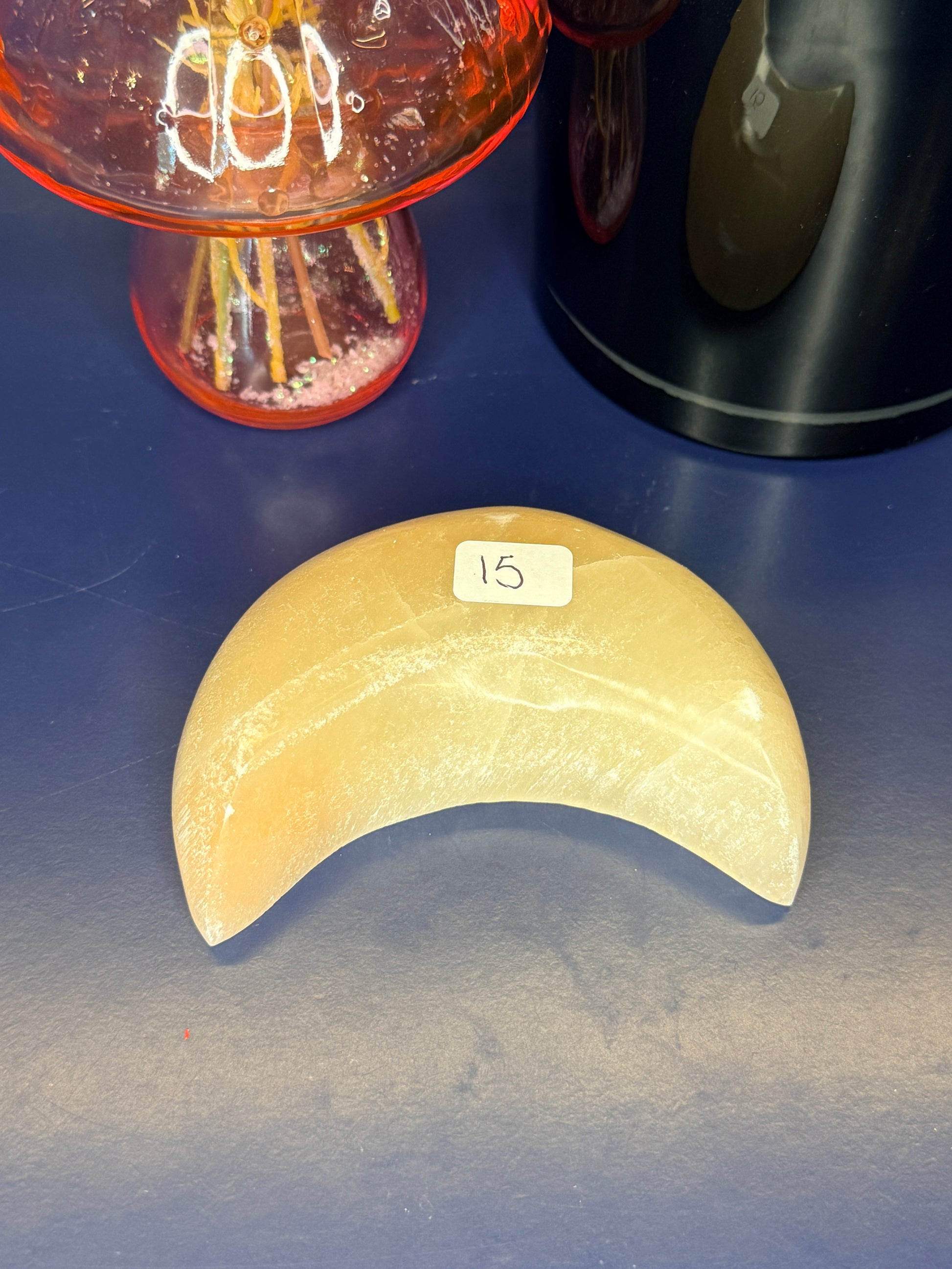 A peach selenite moon bowl with a label sitting in front of a pink mushroom vase and black jar on a blue background