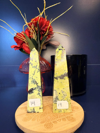 Two lime green opal towers on a wooden coaster in front of a red flower in a vase and black jar