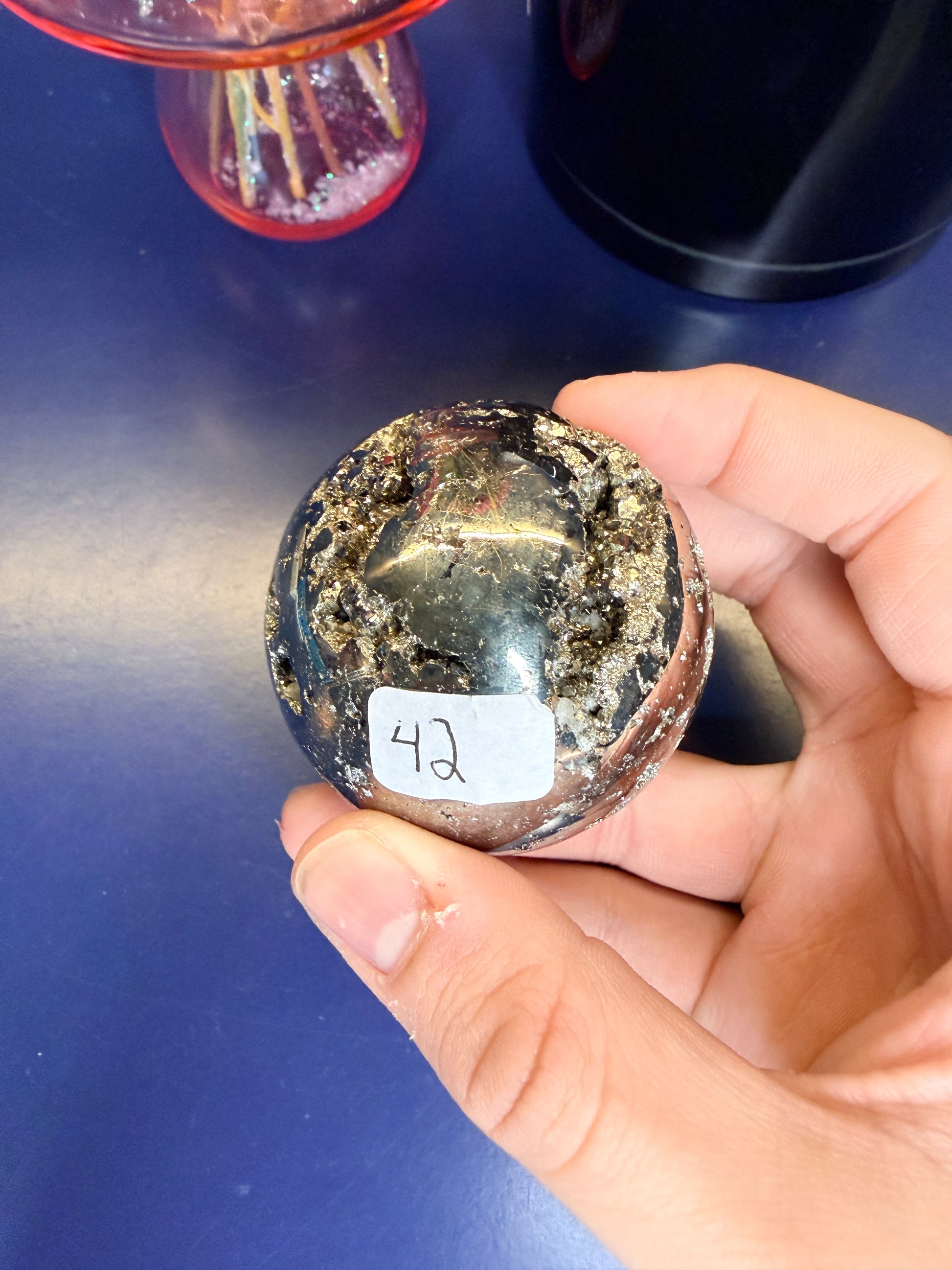 Hand holding a gold colored pyrite sphere in front of a black jar and pink mushroom on a blue background