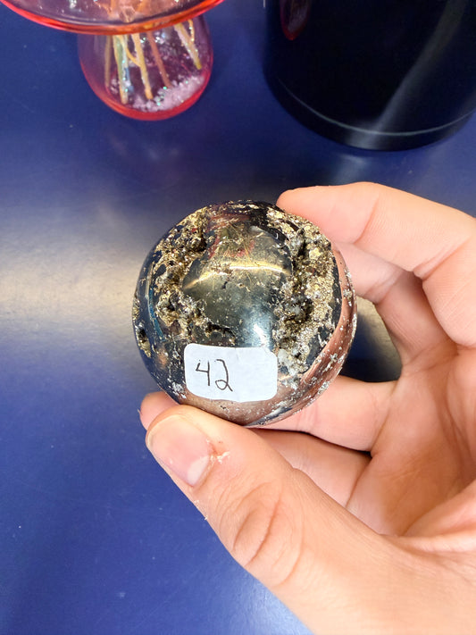 Hand holding a gold colored pyrite sphere in front of a black jar and pink mushroom on a blue background