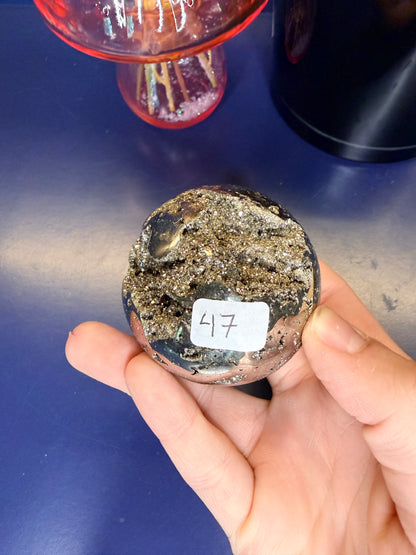 Hand holding a gold colored pyrite sphere in front of a black jar and pink mushroom on a blue background