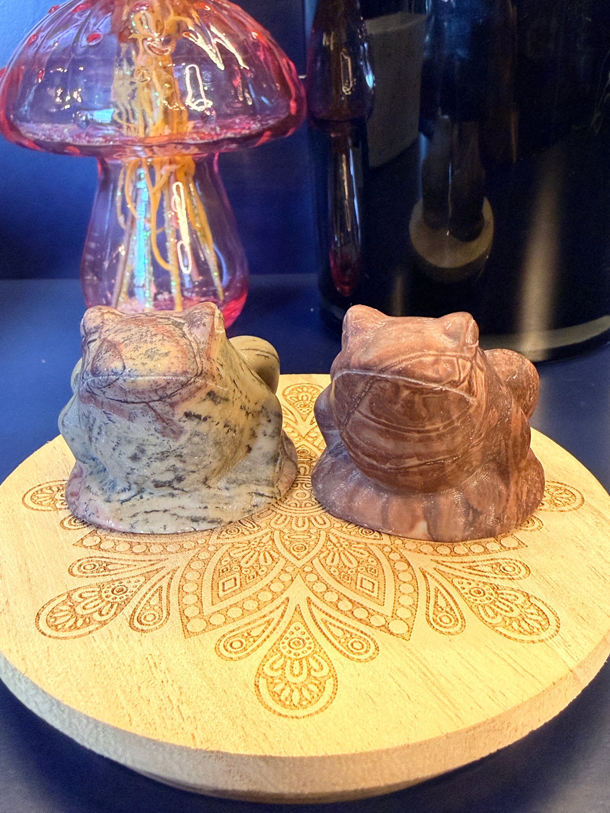 Two large picture jasper frogs sitting on a decorative wooden coaster in front of a glass mushroom and black jar