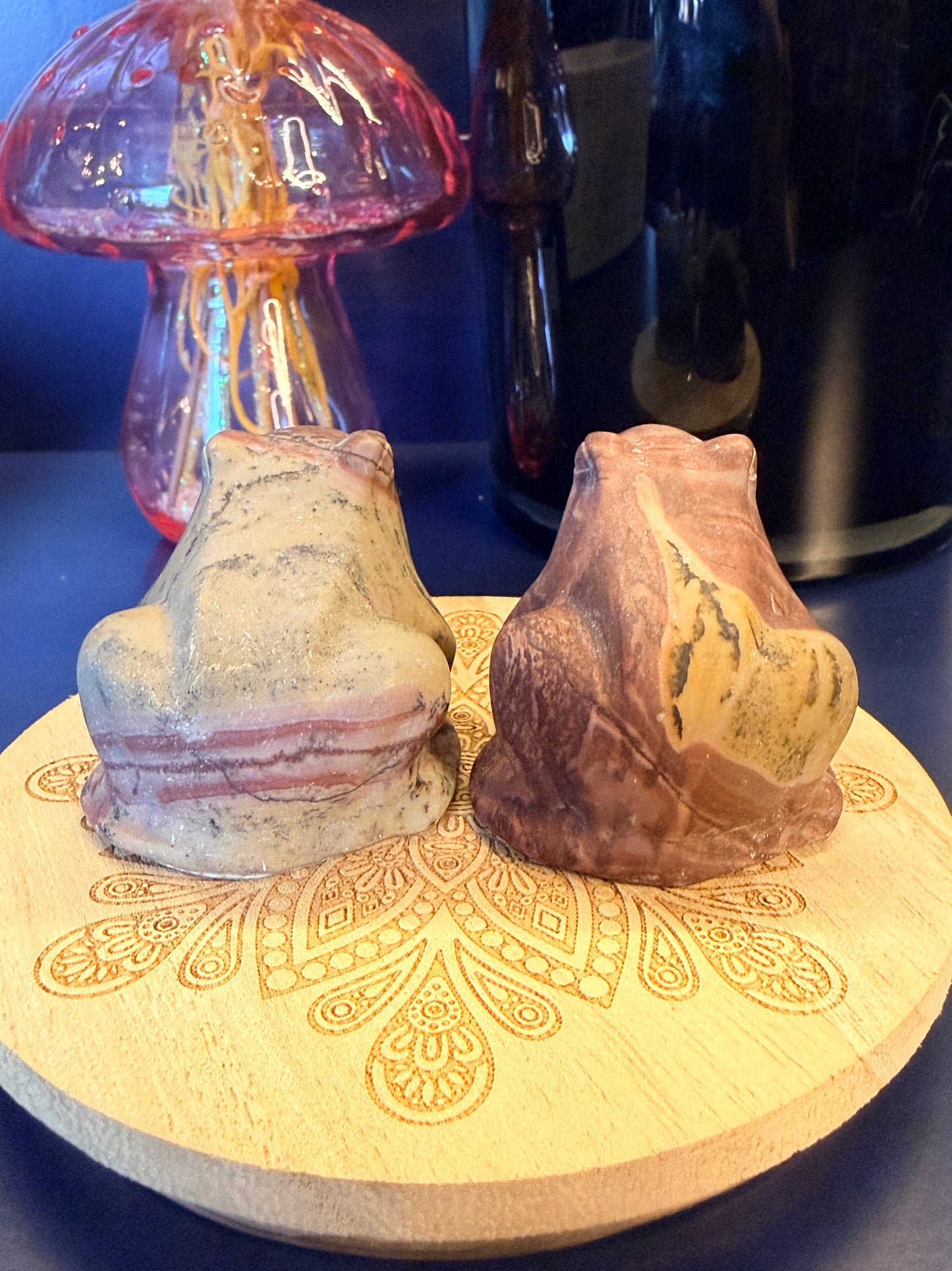 Two large picture jasper frogs sitting on a decorative wooden coaster in front of a glass mushroom and black jar